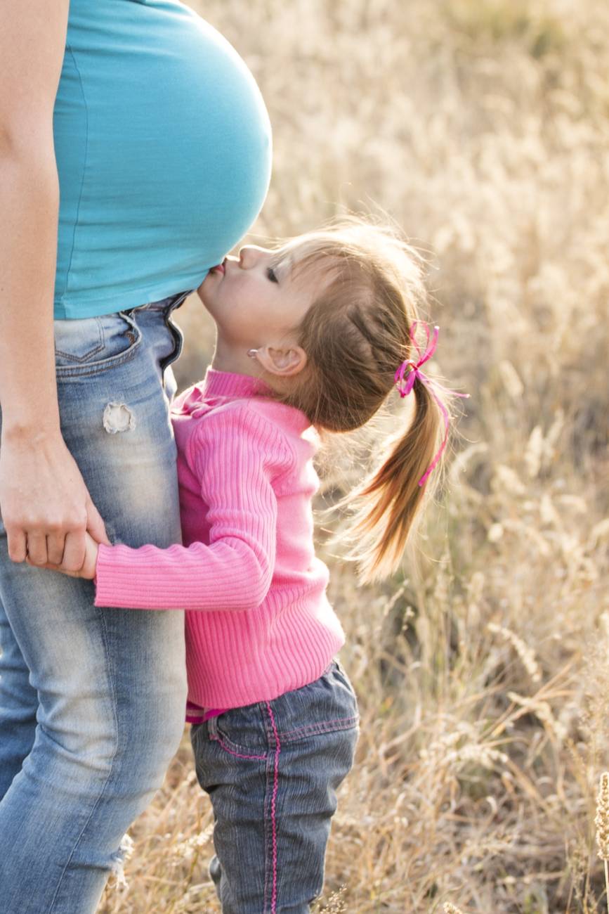 girl in pink sweater and grey jeans kissing tummy of pregnant woman in blue shirt and blue denim jeans