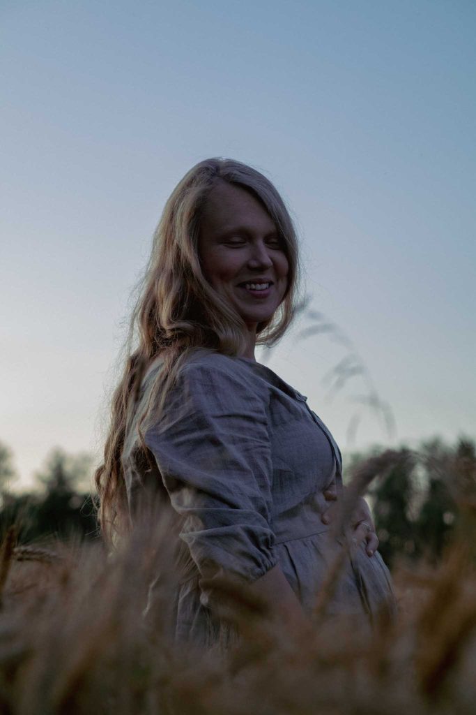 Raskaana oleva nainen seisoo pellolla hymyillen, käsi vatsan päällä, ilta-auringon pehmeässä valossa