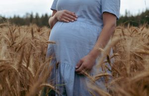 Raskaana oleva nainen seisoo vehnäpellossa käsi vatsan päällä vaaleansinisessä mekossa.