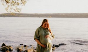 Raskaana oleva nainen seisoo järven rannalla ilta-auringossa ja pitää kättään vatsansa päällä, rauhallisena ja onnellisena alkuraskaudessa.