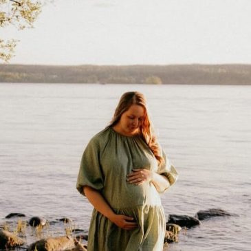 Raskaana oleva nainen seisoo järven rannalla ilta-auringossa ja pitää kättään vatsansa päällä, rauhallisena ja onnellisena alkuraskaudessa.