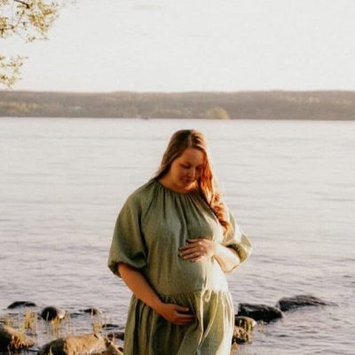 Raskaana oleva nainen seisoo järven rannalla ilta-auringossa ja pitää kättään vatsansa päällä, rauhallisena ja onnellisena alkuraskaudessa.